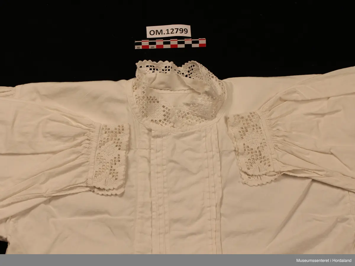 Bunadskjorte i bomullslerret med halskrage og handlinningar i hardangersaum. Brodert på svært tynt bomullstoff. Skjorta har isydde ermer med litt rynker på ermetoppen. Framme på ermene er det og rynker. Knapphol på ermelinngane til mansjettknappar. Midt framme er det ein brei stolpe med biser (legg) på kvar side. Ingen annan dekor på denne. Linningane er sydde på med hand etter at skjorta er heilt ferdig. Det er smale kanteband både framme på ermene og rundt halsen som dekker sårkantane. Så er hardangersaumlinningane nerast tråkla på. Ein rustflekk framme på stolpen.