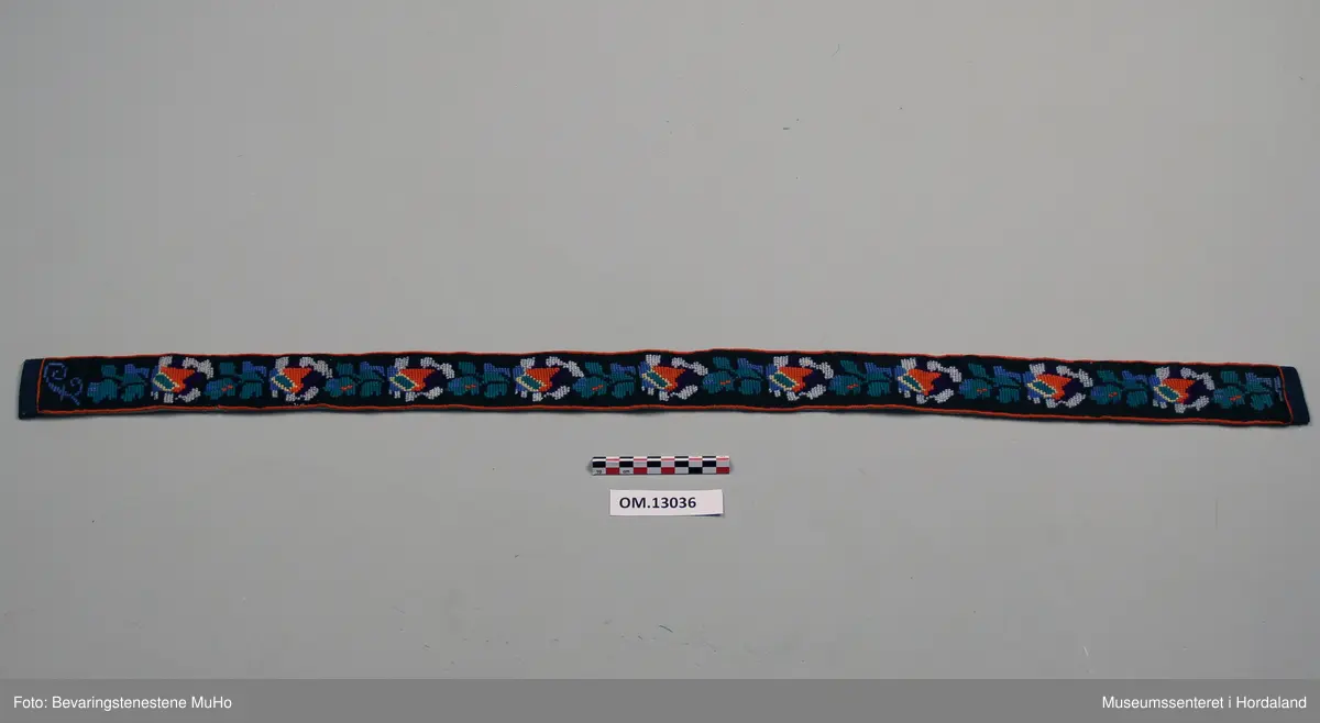 Ei lengd av ein sele brodert med ulltråd. Broderiet har halve krossting sydd i ulike retningar. Svart botnfarge. Kanting er laga ved å sy ein omgang raude skråsting rundt heile selen. Selen er brodert med blomstermønster i mange fargar, og dekorert med lyseblåe perler. I begge kortendane er det sydd på eit mørkeblått kantband som går over på baksida. Bokstaven F er brodert etter blomstermønsteret i eine enden. Kvit- og blåstripete bakstoff i bomull.