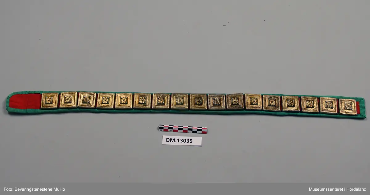 Stølabelte med 17 firkanta sylvstølar med forgylling. Stølane er monterte på raudt ullklede. Beltet er kanta med grøn silkekvarde og er noko avrunda i eine enden. I denne enden er det 8 cm utan påmonterte stølar. Bakstoff er kvitt bomullsstoff.