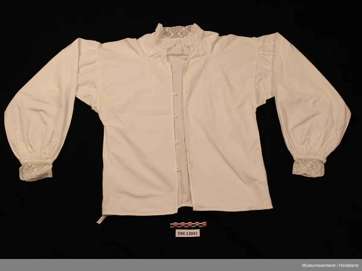Kvit bunadsskjorte i bomull, med hekla kantar. Motivet er åttebladroser. Kile under armane og oppover på skuldrane. Nokre legg heilt øvst på armane og rynking rundt hals og framme på armane. Skjorta er heilt open framme og har hemper av bomullsband og knappar til attlat. På armane er det ein knapp med hempe. Maskinsaumar