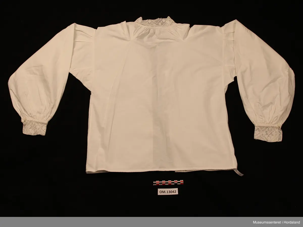Kvit bunadsskjorte i bomull, med hekla kantar. Motivet er åttebladroser. Kile under armane og oppover på skuldrane. Nokre legg heilt øvst på armane og rynking rundt hals og framme på armane. Skjorta er heilt open framme og har hemper av bomullsband og knappar til attlat. På armane er det ein knapp med hempe. Maskinsaumar