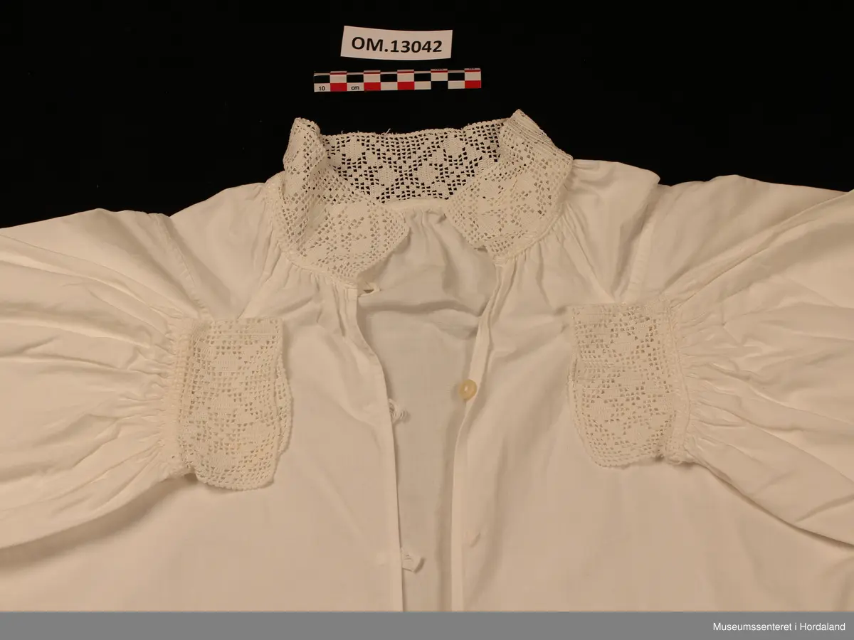 Kvit bunadsskjorte i bomull, med hekla kantar. Motivet er åttebladroser. Kile under armane og oppover på skuldrane. Nokre legg heilt øvst på armane og rynking rundt hals og framme på armane. Skjorta er heilt open framme og har hemper av bomullsband og knappar til attlat. På armane er det ein knapp med hempe. Maskinsaumar