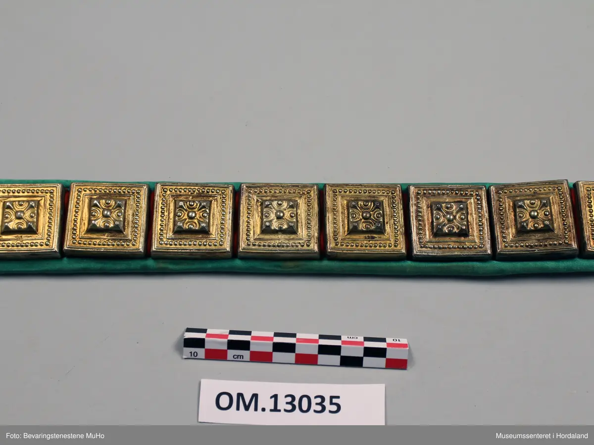 Stølabelte med 17 firkanta sylvstølar med forgylling. Stølane er monterte på raudt ullklede. Beltet er kanta med grøn silkekvarde og er noko avrunda i eine enden. I denne enden er det 8 cm utan påmonterte stølar. Bakstoff er kvitt bomullsstoff.