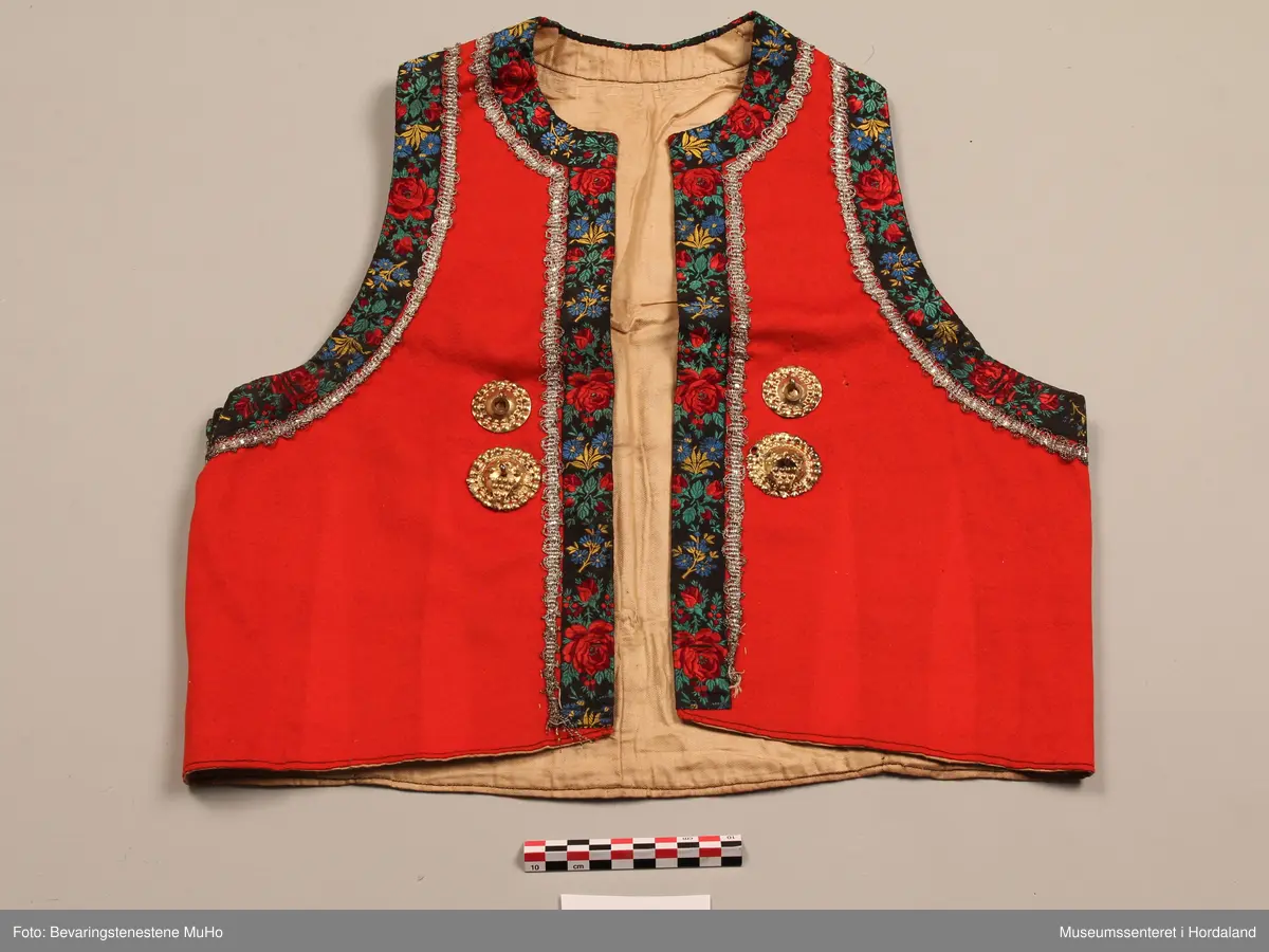 Raudt liv av ull. Kanta med breitt roseband i silke. Innom desse er ein sølvfarga metallknipling. To forgylte borer på kvar side framme. Lysbrunt forstoff i bomull. Fire hekter nede framme.