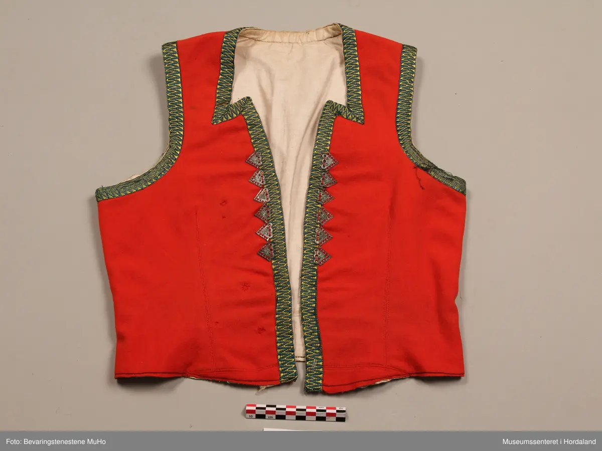 Liv i raudt ullstoff, med grøne og gule krokakvardingar av silke rundt opningane. 6 trekantar av tinn på kvar side av opninga framme. Livet har spissar i bringa. Ubleika lerretfor i bomull. 4 hekter nederst i opninga for attlat. 3 hekter bak på ryggen. Eit legg på kvar side framme med stikningar, og ein ekstra saum på kvar side av ryggen. På eine sida framme er det nokre små hol som er reparert.