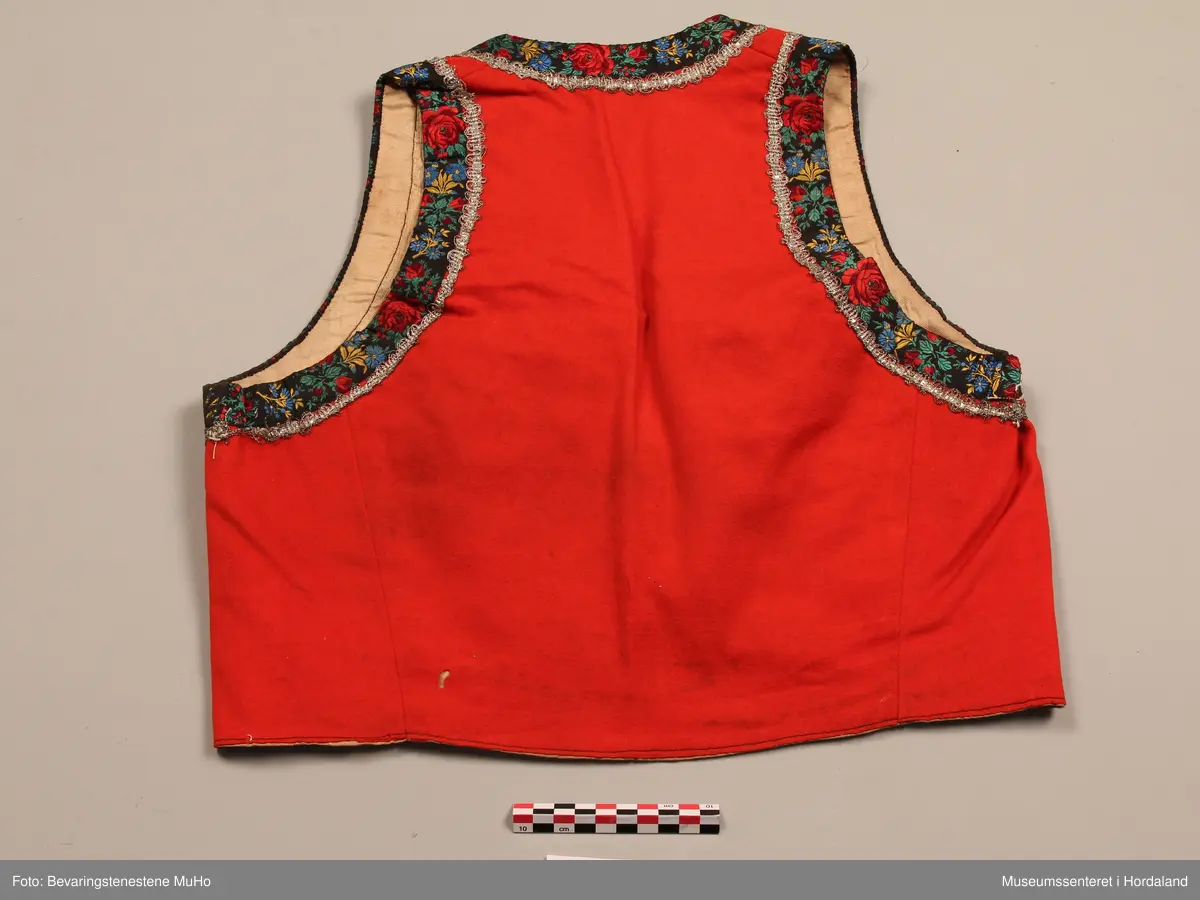 Raudt liv av ull. Kanta med breitt roseband i silke. Innom desse er ein sølvfarga metallknipling. To forgylte borer på kvar side framme. Lysbrunt forstoff i bomull. Fire hekter nede framme.