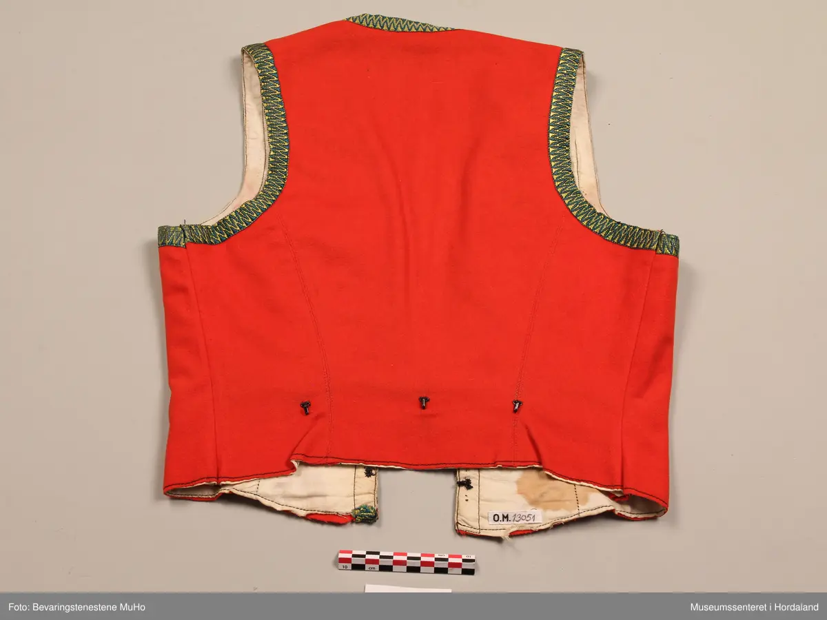 Liv i raudt ullstoff, med grøne og gule krokakvardingar av silke rundt opningane. 6 trekantar av tinn på kvar side av opninga framme. Livet har spissar i bringa. Ubleika lerretfor i bomull. 4 hekter nederst i opninga for attlat. 3 hekter bak på ryggen. Eit legg på kvar side framme med stikningar, og ein ekstra saum på kvar side av ryggen. På eine sida framme er det nokre små hol som er reparert.