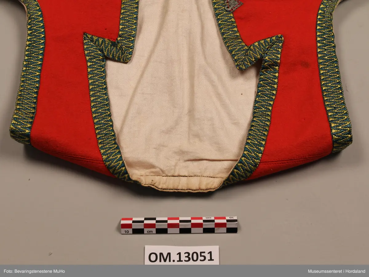 Liv i raudt ullstoff, med grøne og gule krokakvardingar av silke rundt opningane. 6 trekantar av tinn på kvar side av opninga framme. Livet har spissar i bringa. Ubleika lerretfor i bomull. 4 hekter nederst i opninga for attlat. 3 hekter bak på ryggen. Eit legg på kvar side framme med stikningar, og ein ekstra saum på kvar side av ryggen. På eine sida framme er det nokre små hol som er reparert.