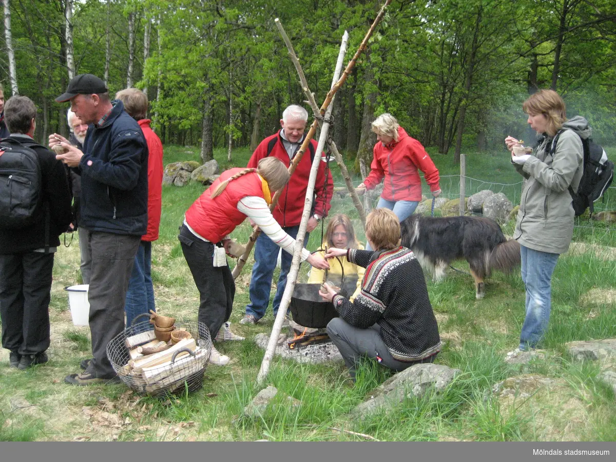 Besökare som deltar vid invigningen av Fagereds natur- och kulturstig i Lindome år 2008. Museitekniker Sven-Åke Svensson, klädd i röd jacka och blåjeans, står och bevakar elden där en stor järngryta hänger i en träställning ovanför. Till vänster ses en korg med ved och handgjorda dryckeskärl av trä. Personer i närheten äter ur små skålar. 
Fagered bjuder på kommunens bäst bevarade järnåldersmiljö. Här finns rester av en gammal hålväg, spår av äldre jordbruksmark och ett 40-tal gravar. 
Relaterade motiv: 2024_1300 - 1308.