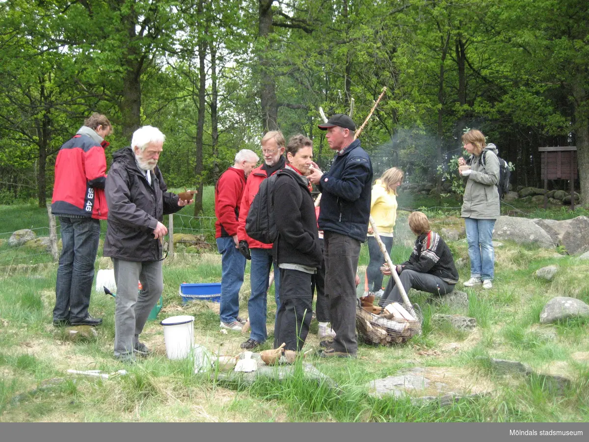 Besökare som deltar vid invigningen av Fagereds natur- och kulturstig i Lindome år 2008. Tredje person från vänster är museitekniker Sven-Åke Svensson, klädd i röd jacka och blåjeans, som står och bevakar elden. På marken ligger/står spänner. Personer i närheten äter ur små skålar. 
Fagered bjuder på kommunens bäst bevarade järnåldersmiljö. Här finns rester av en gammal hålväg, spår av äldre jordbruksmark och ett 40-tal gravar. 
Relaterade motiv: 2024_1300 - 1308.
