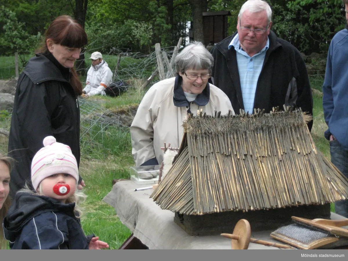 Besökare deltar vid invigningen av Fagereds natur- och kulturstig i Lindome år 2008. Ett litet barn sitter på knä framför en stenåldersby i mini-format (se 1988_0229). Byn består av ett hus, ställningar för att torka hud samt trädjur. Närmast barnet står museichef Mari-Louise Olsson (klädd i svart). I närheten står fler vuxna och beundrar den lilla byn. 
Fagered bjuder på kommunens bäst bevarade järnåldersmiljö. Här finns rester av en gammal hålväg, spår av äldre jordbruksmark och ett 40-tal gravar. 
Relaterade motiv: 2024_1300 - 1308.