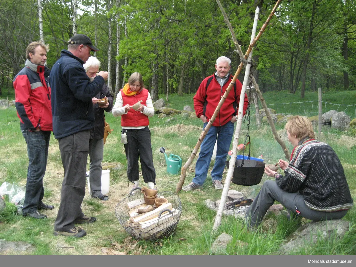 Besökare som deltar vid invigningen av Fagereds natur- och kulturstig i Lindome år 2008. Museitekniker Sven-Åke Svensson, klädd i röd jacka och blåjeans, står och bevakar elden där en stor järngryta hänger i en träställning ovanför. Till vänster ses en korg med ved och handgjorda dryckeskärl av trä. Personer i närheten äter ur små skålar. 
Fagered bjuder på kommunens bäst bevarade järnåldersmiljö. Här finns rester av en gammal hålväg, spår av äldre jordbruksmark och ett 40-tal gravar. 
Relaterade motiv: 2024_1300 - 1308.