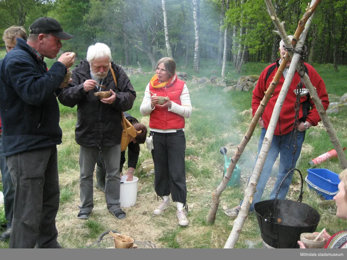 Besökare som deltar vid invigningen av Fagereds natur- och kulturstig i Lindome år 2008. Museitekniker Sven-Åke Svensson, klädd i röd jacka och blåjeans, står och bevakar elden där en stor järngryta hänger i en träställning ovanför. På flera ställen står spänner med vatten. Personer i närheten äter ur små skålar och längst ner ses handgjorda dryckeskärl av trä. 
Fagered bjuder på kommunens bäst bevarade järnåldersmiljö. Här finns rester av en gammal hålväg, spår av äldre jordbruksmark och ett 40-tal gravar. 
Relaterade motiv: 2024_1300 - 1308.