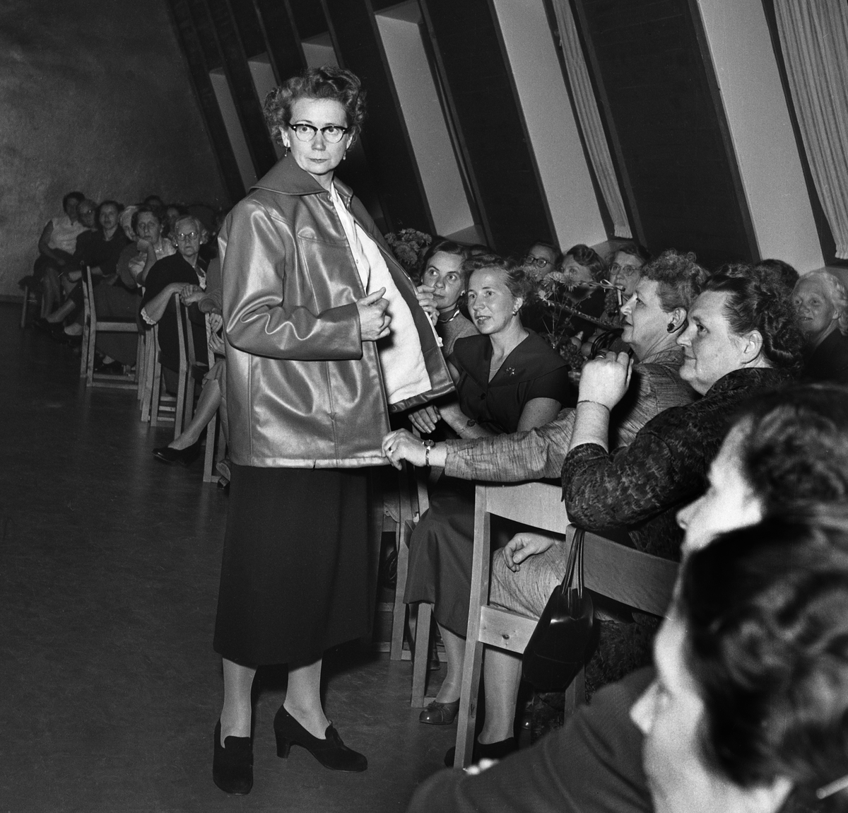 Mannekänguppvisning i Linköping.
Modevisningen är på Åbylundstorget 2 i en samlingslokal. Kvinnan visar en jacka.


...

385 bilder om Linköping på 1950-talet från tidningen Östgötens arkiv. Framtidstro och optimism är ord som sammanfattar Linköping på femtiotalet. Årtiondet innebar satsningar för att förbättra linköpingsbornas livsvillkor. Bostadsfrågan och trafiklösningarna dominerade den lokalpolitiska agendan.
Bilderna digitaliserades år 2013.