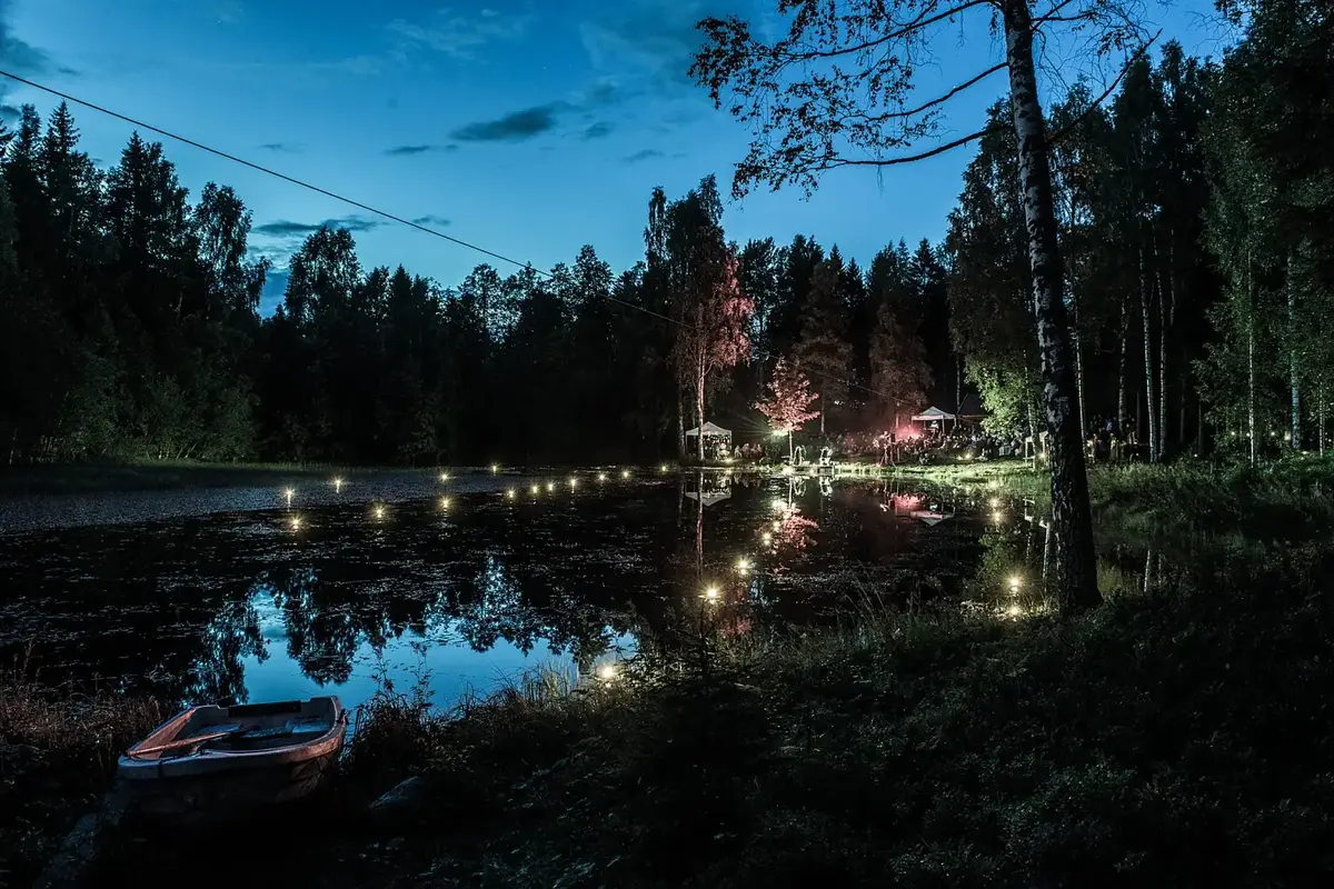 Et tjern i skogen rett etter solnedgang. En opplyst enkel scene i den andre enden av vannet, og flytende fakler. 