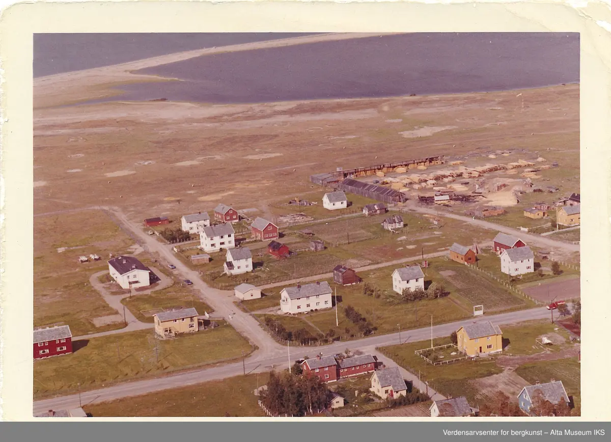Oversiktsbilde fra Widerøe av Elvebakken, med utsikt over bolighus, et sagbruk, uthus og jorder som står langs Altaveien. 