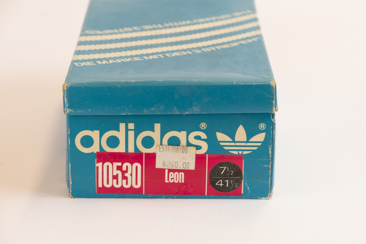 Blå skoeske inneholdende et par svarte fotballsko og en blå knottenøkkel.

Esken er blå med hvit skrift og logo på hver side med tilhørende løst lokk. På den ene siden er festet en rosa lapp med informasjon og en hvit prislapp.

Skoene er svarte med hvite detaljer, naturell gummisåle med røde skruknotter på undersiden. 

En blå knottenøkkel i plast. En hette med vinger som stikker opp og ut til å ta tak med.