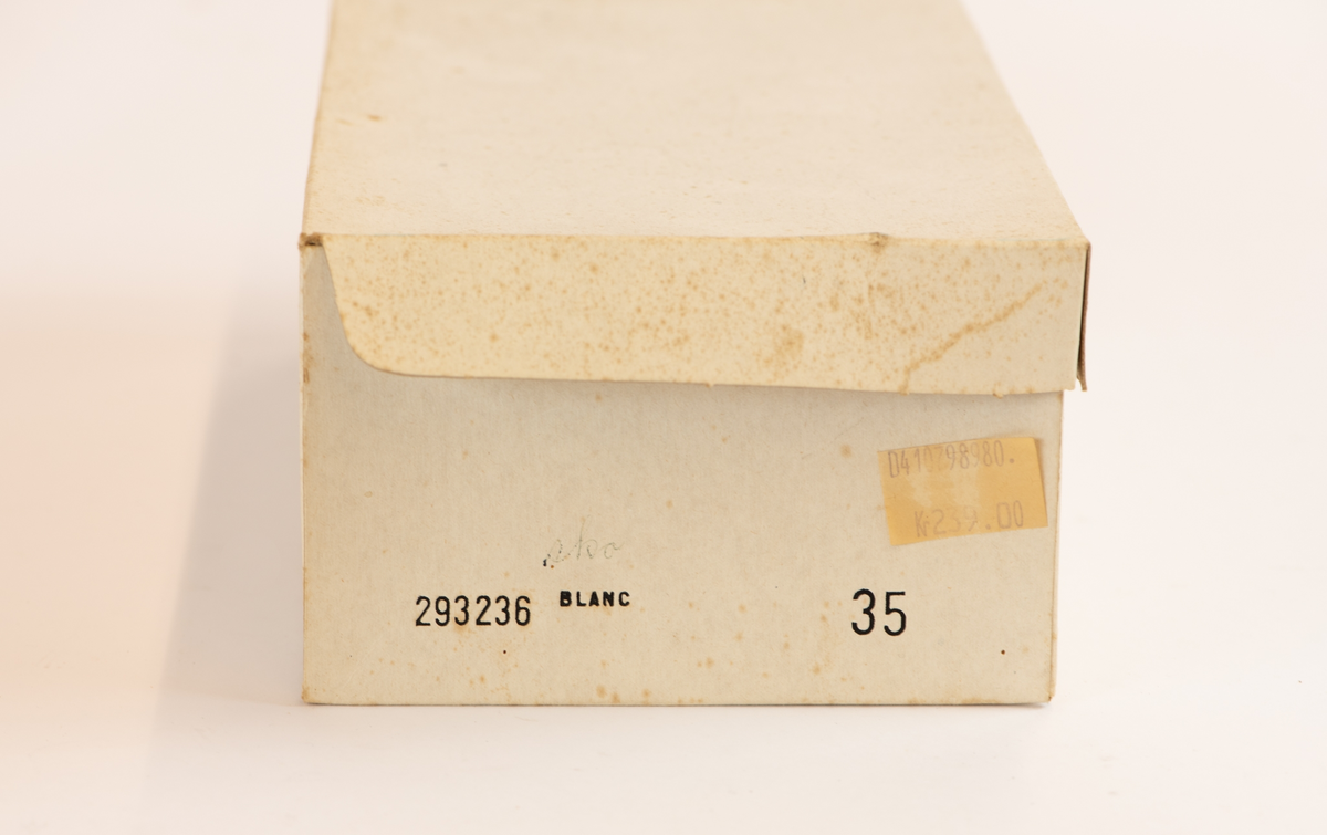 Mokkasiner i originaleske. Str. 35. Hvite, med brun såle. Frynser foran, disse er spisset i enden og har et lite hull. Et felt med innfelt rutete stoff, midt i dette knyttet en frynse. Ligger i kombinert eske/lokk.