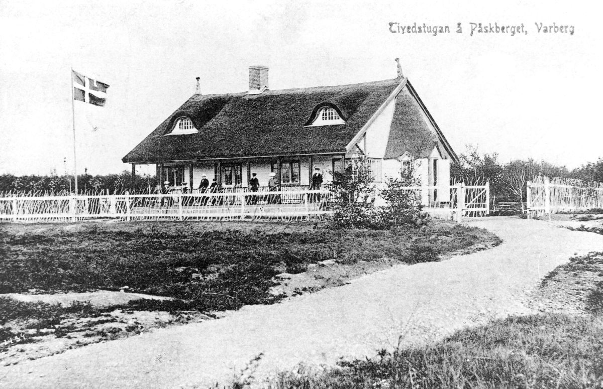 Vykort, "Tivedstugan å Påskberget, Varberg." Serveringsbyggnaden uppfördes 1906 i Påskbergsskogen och ritades av Gottfrid Ljunggren. Namnet kommer sig troligen av att han kom från Töreboda, där en del av Tiveden ligger. Där gavs fri bostad och fri ved utöver lönen till Påskbergets vaktmästare. Den förste hette Karl Nilsson och anställdes i juni 1906. En samlingssal inrymdes också i huset med servering under sommarhalvåret. Ett vitmålat staket, delvis bestående av tunna trädgrenar, omger huset och till vänster i bild vajar flaggan på stång.