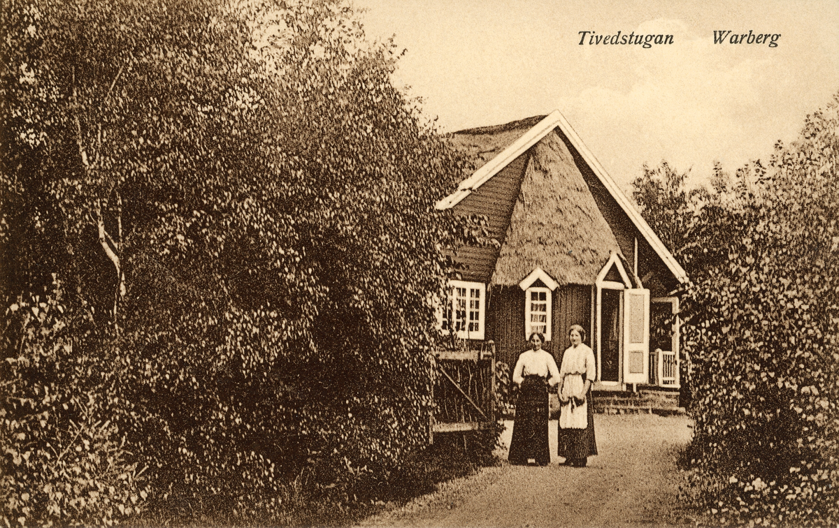 Vykort, "Tivedstugan - Warberg." Två kvinnor står vid grinden in till Tivedstugan i Påskbergsskogen. Där inrymdes både vaktmästarbostad och servering. Dörren står öppen på gavelns ovanliga "absid" med någon form av stråtäckning på taket. Byggnaden lär vara ritad av byggnadsingenjör Gottfrid Ljunggren, arkitekt bakom många byggnader i Varberg 1903-1914.