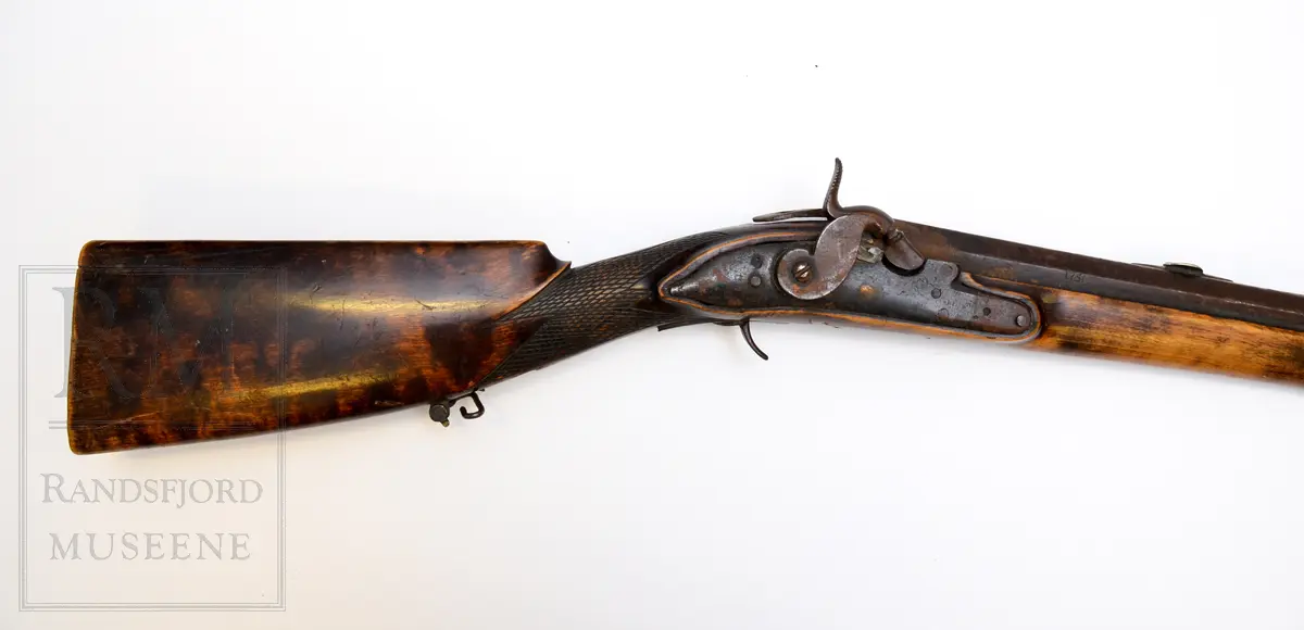 Munnladning, glattløpet m/ perkusjonslås. Skjefte av tre, brunbeiset. Kolben har kinntre og utskjæringer (nettskjæring) på kolbehalsen. Pipe og lås av jern m/ fram og baksikte i messing. To rembøyler med skruer. Ladestokk (b)  av tre er festet under forskjeftet. Bøylebeslag, avtrekkerbøyle mangler. gjennomgående skrue mellom beslag bak pipe og avtrekkerbeslag mangler. Forskjeftet går helt fram til pipemunningen. Har ikke bajonett. Pipen er rund framme og åttekantet bak. Grovkalibret.