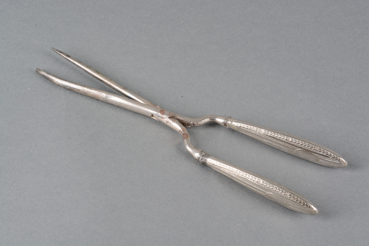 Tynn krølltang med håndtak i metall, med preget dekor på håndtakene. Det er dekorert med Louis XVI-mønster i lavt relieff: damask-striper med rose- og bladfrise langs kanten og rundt et ovalt felt for monogram (det er ikke gravert noe monogram). Det er mønster på begge sidene av skaftet. Spor av varmebehandling. på selve tangen.