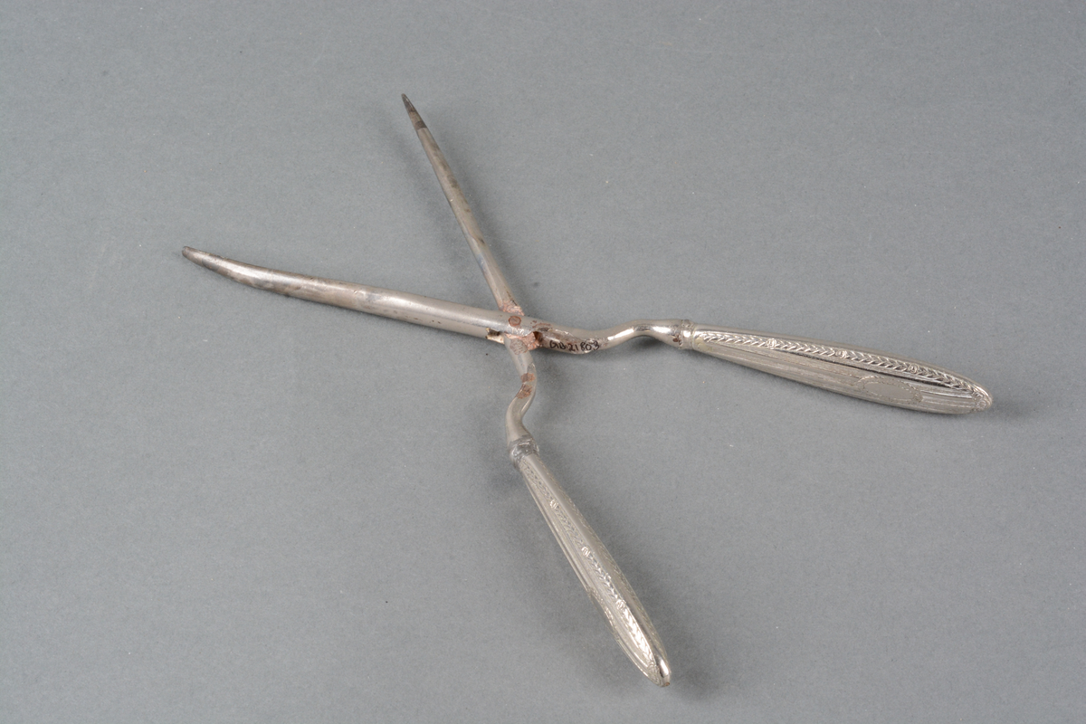 Tynn krølltang med håndtak i metall, med preget dekor på håndtakene. Det er dekorert med Louis XVI-mønster i lavt relieff: damask-striper med rose- og bladfrise langs kanten og rundt et ovalt felt for monogram (det er ikke gravert noe monogram). Det er mønster på begge sidene av skaftet. Spor av varmebehandling. på selve tangen.