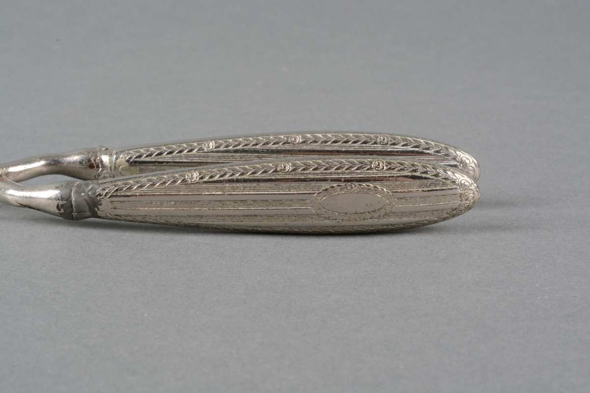 Tynn krølltang med håndtak i metall, med preget dekor på håndtakene. Det er dekorert med Louis XVI-mønster i lavt relieff: damask-striper med rose- og bladfrise langs kanten og rundt et ovalt felt for monogram (det er ikke gravert noe monogram). Det er mønster på begge sidene av skaftet. Spor av varmebehandling. på selve tangen.
