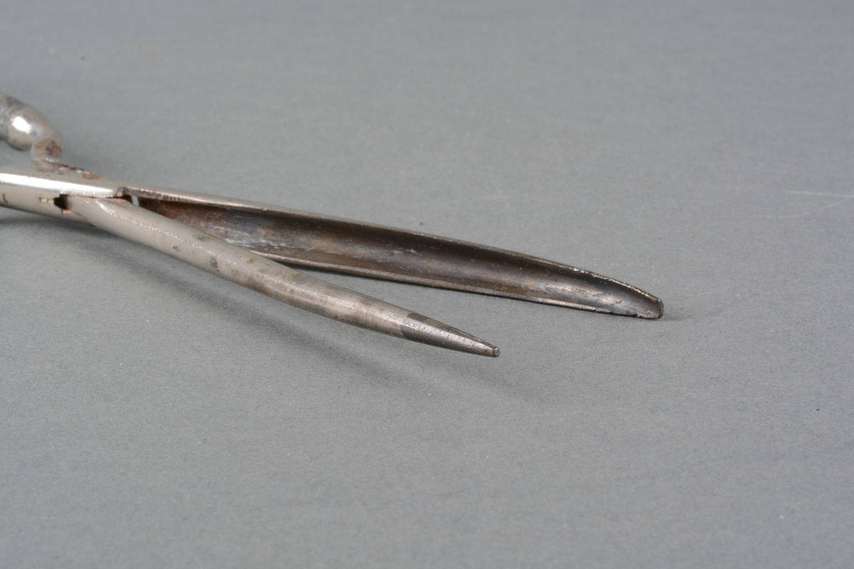 Tynn krølltang med håndtak i metall, med preget dekor på håndtakene. Det er dekorert med Louis XVI-mønster i lavt relieff: damask-striper med rose- og bladfrise langs kanten og rundt et ovalt felt for monogram (det er ikke gravert noe monogram). Det er mønster på begge sidene av skaftet. Spor av varmebehandling. på selve tangen.