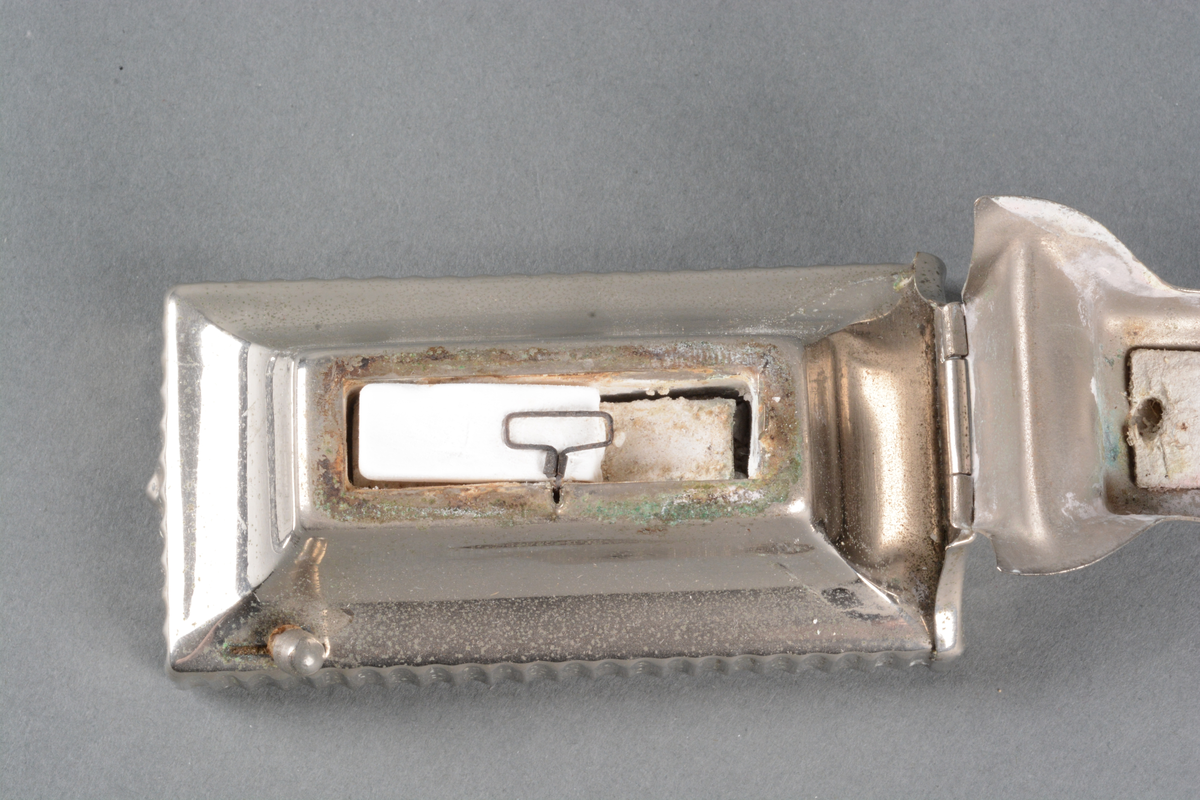 Krølltangvarmer med metatablett, laget av forniklet metallplate med hengslet lokk som fungerer som holder for en krølltang. Det ligger en metatablett i.
