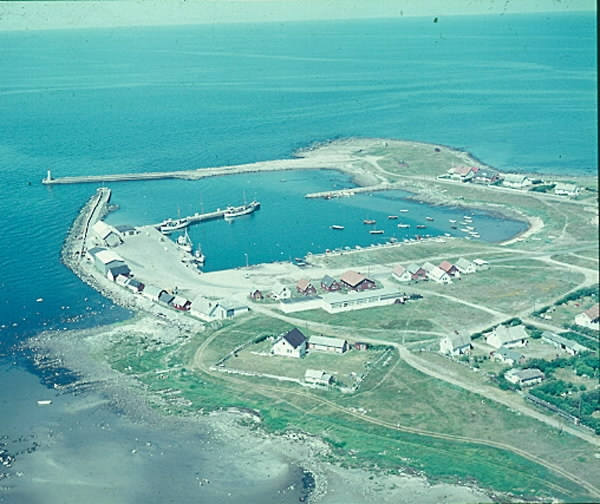 Flygbild. Glommens hamn med fiskebåtar och fritidsbåtar, magasin, sjöbodar och omgivande bostadsbebyggelse. Ute på näset står Glumstenen, flyttblocket från istiden som gett fiskeläget Glommen sitt namn, anlagt på 1880-talet. Till en början kallades då både stenen och platsen för Glummen. Stenen har varit ett landmärke för sjöfarare i alla tider. I isländska skrifter benämns den som Glaumstein. Ordet "glum" stammar från något av de fornnordiska orden glaumr, glumr som betyder larma, bullra. Förr användes ordet "bullersten" om flyttblock (på engelska "boulder"), vilket också har koppling till ordet "bumling".