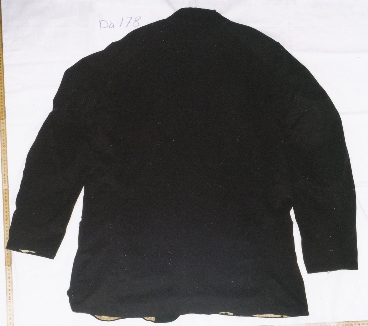 Kostym, brudgumskostym, bestående av kavaj, långbyxor och väst i svart tuskaftad ylle, enligt uppgift är tyget hemvävt. Handsydd av skräddare.
a. Kavaj. Två framstycken/sidstycken med tillbakadragna axelsömmar, smal nedliggande krage och slag, enkelknäppt med tre präglade svarta läderknappar och knapphål tränsade med svart sysilke, framkanterna något rundade nertill, två snedställda stolpfickor och en snedställd bröstficka på vänster sida. Helt ryggstycke med svängda ryggsömmar, isydd ensömsärm. Dekorsöm vid nederkanten och på bröstfickan med vågig efterstygnssöm. Foder i bålen av tryckt bomullstyg med små ovaler i beige och brunt, innerficka på vänster sida, ärmarna fodrade med grov vit linnelärft. 

b. Byxor. Två framstycken, två bakstycken förhöjda i midjan, ingen linning, två fickor i sidsömmen, dold gylfknäppning med tre knappar, knapphålen sydda med brunt lingarn, fyra gråbruna hängselknappar av ben. Helfodrade med grov vit linnelärft.

c. Väst. Framstycken och krage i ylletyget, tillbakadragna axelsömmar, liten nedliggande krage med rundade hörn, dubbelknäppt med sex par gråbruna benknappar, två extra knappar att knäppa ut slagen med, endast det vänstra slaget är infodrat med det svarta tyget, två stolpfickor. Ett bakstycke av fin vit linnelärft något uppsvängt bak, två spänntampar från sidsömmen med metallspänne. Framstyckena fodrade med hemvävt linnelärft tvärrandat i brunt och beige. Dekorsöm vid nederkanten med vågig efterstygnssöm.

Anteckning: Brudgumskostym buren av Karl Gustaf Karlsson, Kroken, Skårtaryd vid bröllopet 1867. Skänkt av hans barn Hilda, Oskar, Alfred och Edit Karlsson 1939, vilka alla förblev ogifta. Fadern var kyrkvärd i Dädesjö i 30 år.

Birgitta Blixt 2021.