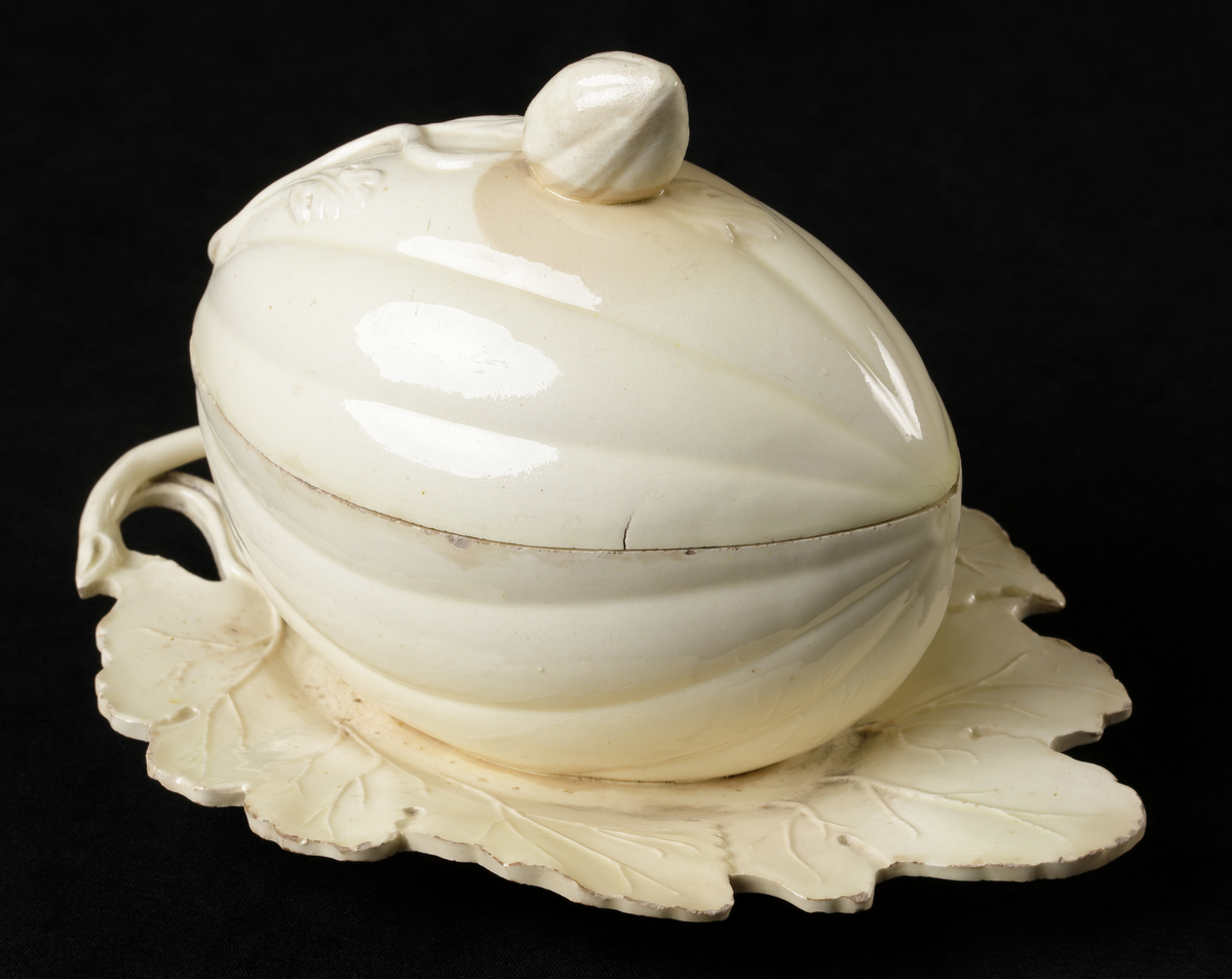 En naturalistiskt formgiven skål med lock, i gulvitt glaserat porslin, utformad som en liten oval pumpa som vilar på ett blad.
Utskårat lock (för sked?).