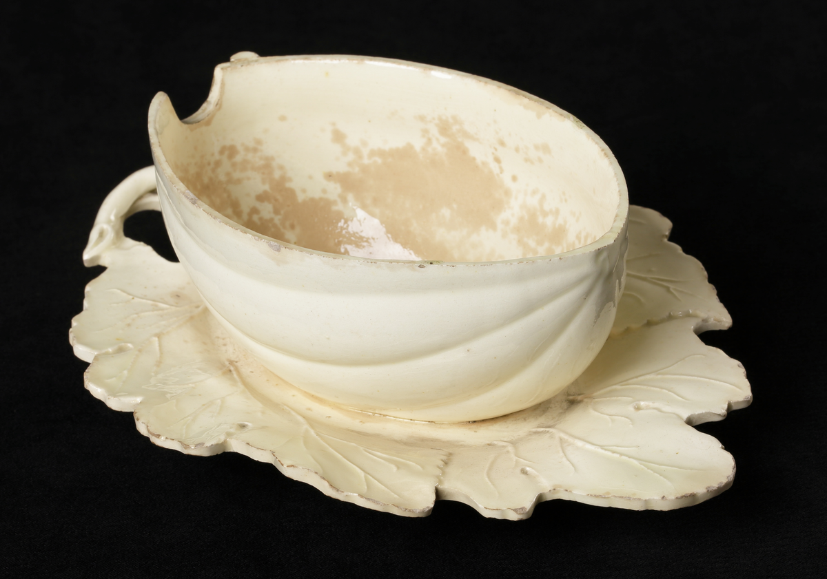 En naturalistiskt formgiven skål med lock, i gulvitt glaserat porslin, utformad som en liten oval pumpa som vilar på ett blad.
Utskårat lock (för sked?).