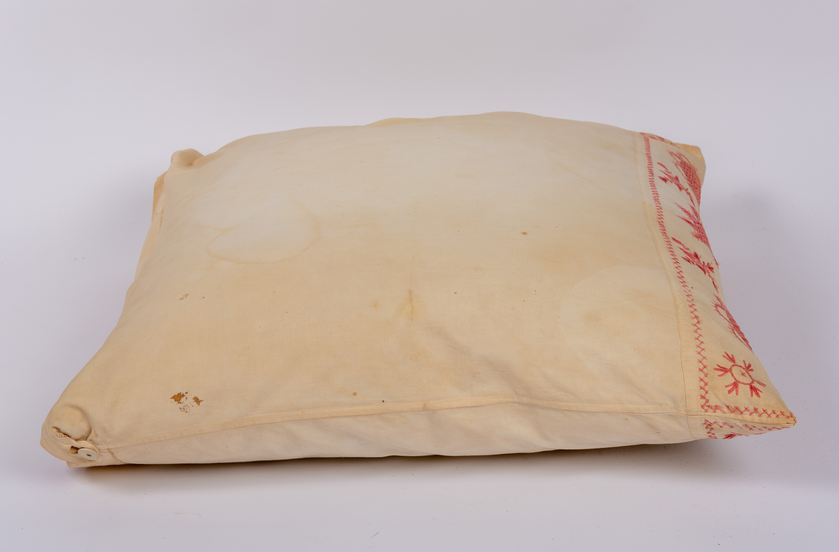 Örngott av vävt bomullstyg med hallandssöm i rött garn samt öppning med klädda knappar och fåll med hålsöm. Innerkudde av rosafärgat modernare tyg.