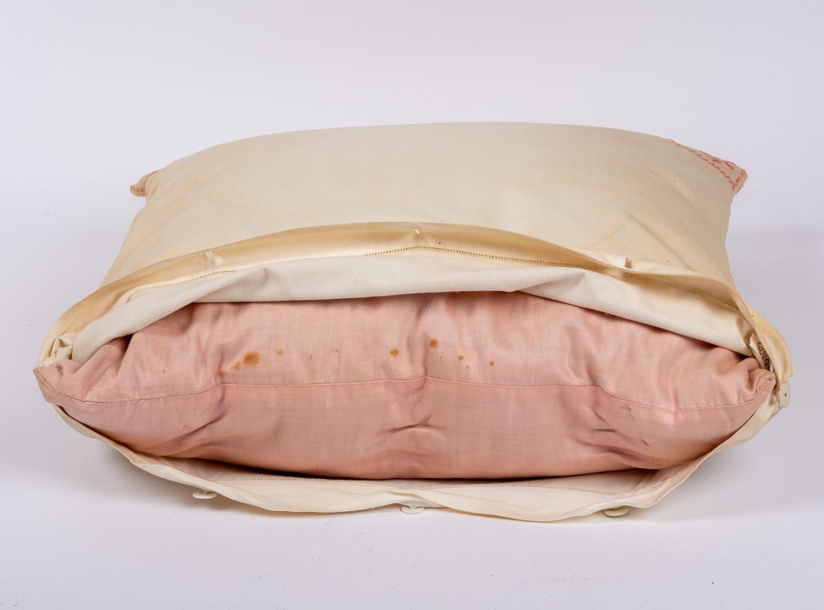 Örngott av vävt bomullstyg med hallandssöm i rött garn samt öppning med klädda knappar och fåll med hålsöm. Innerkudde av rosafärgat modernare tyg.