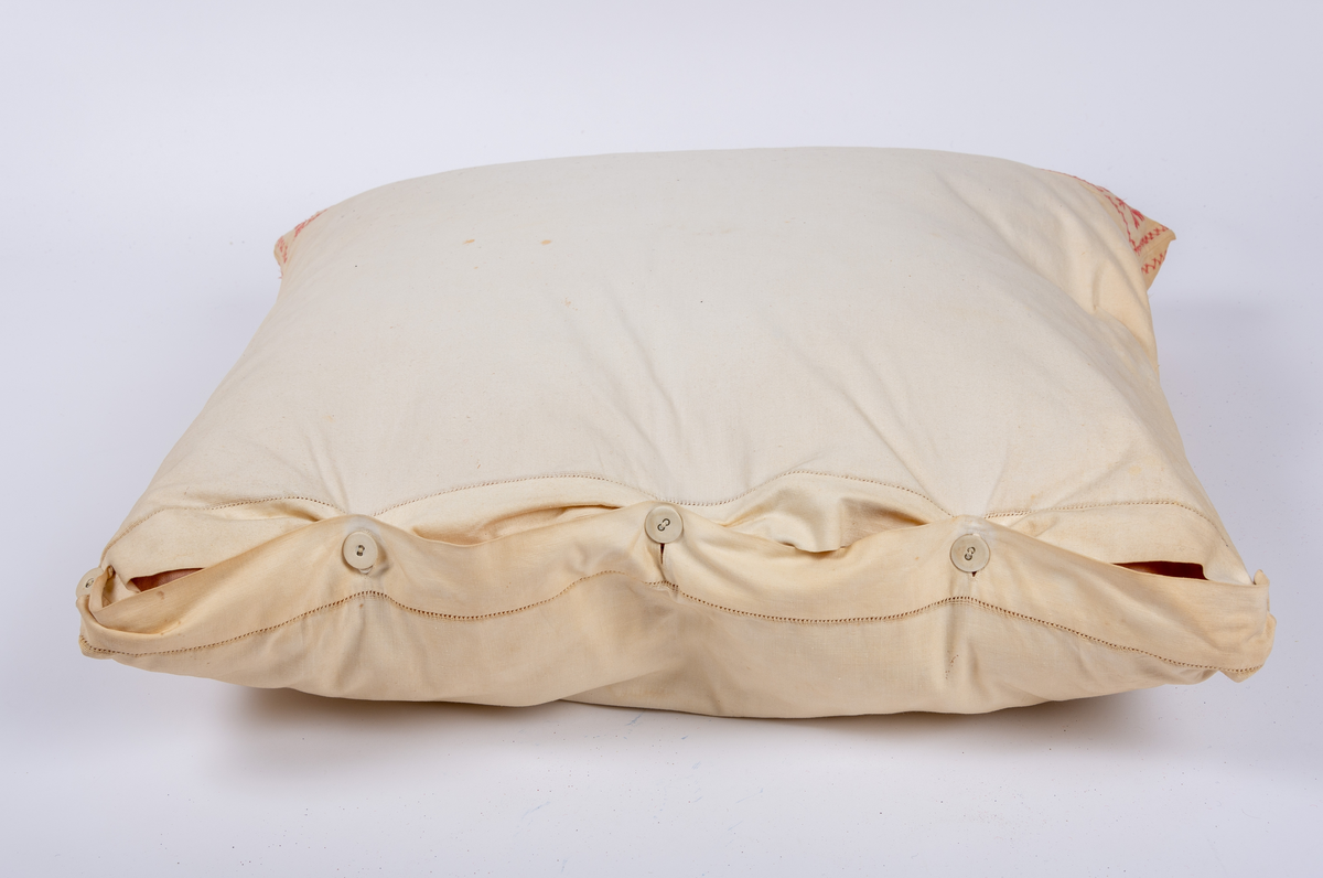 Örngott av vävt bomullstyg med hallandssöm i rött garn samt öppning med klädda knappar och fåll med hålsöm. Innerkudde av rosafärgat modernare tyg.