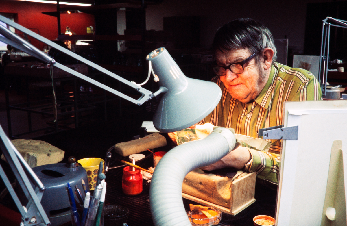 Gravyrverkstaden på ett glasbruk. En man med glasögon sitter och graverar.
Färgfoto, ca 1973.