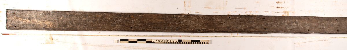 Däcksplanka.
Ena änden är snedskuren.
34 genomgående spikhål. Spikhålen är original från när skeppet byggdes. Originalspikarna är bortrostade. Efter bärgningen monterades däcksplankan med rostfri fransk träskruv samt bricka. Vid demonteringen 2023 lossades alla skruvar. De brickor som gick att ta bort togs bort. En del satt så djupt i träet att det inte gick att demontera dem utan att skada plankan. Dessa brickor fick därför sitta kvar. 
En trädymling i ena änden.
Tydliga avtryck från balkar och ribbor på undersidan som även är täckt av ytliga sprickor och har fläckar av rost.