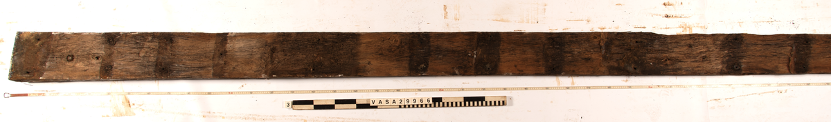 Däcksplanka.
Ena änden är snedskuren.
34 genomgående spikhål. Spikhålen är original från när skeppet byggdes. Originalspikarna är bortrostade. Efter bärgningen monterades däcksplankan med rostfri fransk träskruv samt bricka. Vid demonteringen 2023 lossades alla skruvar. De brickor som gick att ta bort togs bort. En del satt så djupt i träet att det inte gick att demontera dem utan att skada plankan. Dessa brickor fick därför sitta kvar. 
En trädymling i ena änden.
Tydliga avtryck från balkar och ribbor på undersidan som även är täckt av ytliga sprickor och har fläckar av rost.