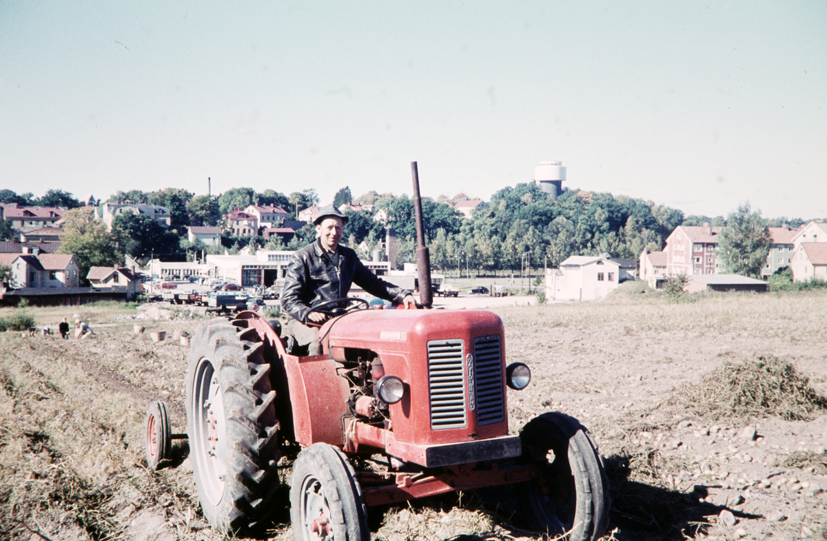 Potatisupplockning på Hov, Växjö. En man kör traktor på den nyupptagna åkern. I bakgrunden ser man kvarteren Mörner, Dalen (numera Lågan) och Lekamensgärdet och det nya vattentornet på Hovsberg. 
Färgfoto ca 1960.