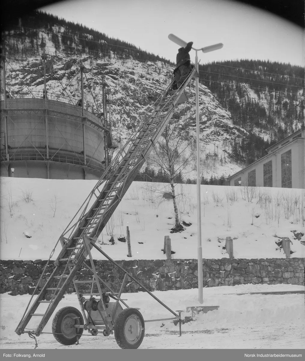 Utskifting av lyspære i lysmast vest for NH3-tapperiet. Mann stående i stige på hjul. N2-gasometer og renseanlgegg i bakgrunnen.