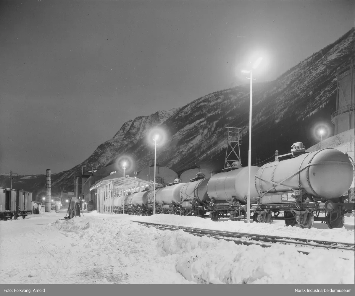 Tankvogner type Q4 stående ved NH3-tapperiet. Belysning langs tapperiets spor. N2-gasometer og Ovnshus 1 i bakgrunnen. To lukkede godsvogner, type G3 på sidespor.