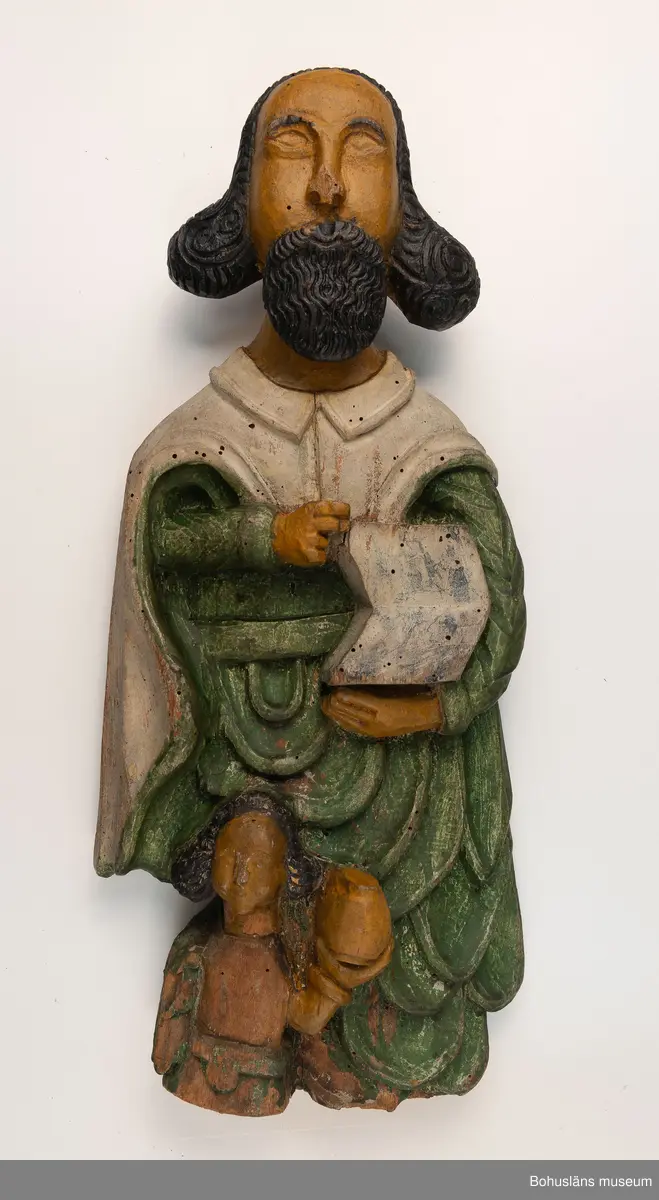 Evangelisten Matteus, nischbild från predikstol. Vid fötterna en ängel. Skägg och hår kraftigt accentuerade. 
Från okänd kyrka i Bohuslän. 
Nischbild från predikstol tillsammans med skulptur UM000399.

G:14 i 1869 års tryckta katalog:
Träbeläte af S:t Matheus.

Knut Adrian Andersosns Katalog I De kyrkliga samlingarna I:124:16:
Träbild av Evangelisten Matheus
44 x 20 cm = G:15 i 1869 års rycka katalog. 
Från tiden efter 1550 i Bohuslän.,

Ur handskrivna katalogen 1957-1958:
S:t Matteus
H.44 cm; i händerna en uppslagen bok, vid fötterna en ängel m. en kalk; manteln grön o. vit, maskhål, färgen ngt. flagn.

Ur Nationalencyklopedin, NE.se: Evangelist
evangelist, benämning på den som enligt traditionen författat något av Nya Testamentets evangelier: Matteus, Markus, Lukas resp. Johannes
I kyrklig bildkonst framställs Matteus och Johannes ofta som äldre män, Markus och Lukas som yngre. Sedan slutet av 300-talet brukar fyra (vanligen bevingade) gestalter, evangelistsymboler, höra ihop med de fyra evangelisterna, antingen bredvid dem eller ensamma, ofta omgivande Kristus eller Lammet. Matteus representeras av en människa, Markus av ett lejon, Lukas av en tjur och Johannes av en örn. Besläktad symbolik möter i tetramorfen ("den fyrgestaltade"), ett bevingat väsen med fyra huvuden (av människa, lejon, tjur och örn), ibland också med motsvarande fötter. Symbolerna är hämtade ur Bibeln. I Hes. 1 skildras hur Hesekiel ser fyra varelser (tetramorfer) kring Guds tronvagn. I Johannes vision i Upp. 4:6-7 har de fyra varelserna kring tronen endast ett ansikte vardera: ett lejon-, tjur-, människo- eller örnansikte. Tillämpningen på evangelisterna bygger på en allegorisk tolkning av Upp. 4:6-7, belagd redan hos Irenaeus.