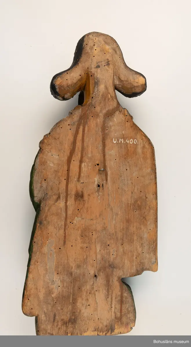 Evangelisten Matteus, nischbild från predikstol. Vid fötterna en ängel. Skägg och hår kraftigt accentuerade. 
Från okänd kyrka i Bohuslän. 
Nischbild från predikstol tillsammans med skulptur UM000399.

G:14 i 1869 års tryckta katalog:
Träbeläte af S:t Matheus.

Knut Adrian Andersosns Katalog I De kyrkliga samlingarna I:124:16:
Träbild av Evangelisten Matheus
44 x 20 cm = G:15 i 1869 års rycka katalog. 
Från tiden efter 1550 i Bohuslän.,

Ur handskrivna katalogen 1957-1958:
S:t Matteus
H.44 cm; i händerna en uppslagen bok, vid fötterna en ängel m. en kalk; manteln grön o. vit, maskhål, färgen ngt. flagn.

Ur Nationalencyklopedin, NE.se: Evangelist
evangelist, benämning på den som enligt traditionen författat något av Nya Testamentets evangelier: Matteus, Markus, Lukas resp. Johannes
I kyrklig bildkonst framställs Matteus och Johannes ofta som äldre män, Markus och Lukas som yngre. Sedan slutet av 300-talet brukar fyra (vanligen bevingade) gestalter, evangelistsymboler, höra ihop med de fyra evangelisterna, antingen bredvid dem eller ensamma, ofta omgivande Kristus eller Lammet. Matteus representeras av en människa, Markus av ett lejon, Lukas av en tjur och Johannes av en örn. Besläktad symbolik möter i tetramorfen ("den fyrgestaltade"), ett bevingat väsen med fyra huvuden (av människa, lejon, tjur och örn), ibland också med motsvarande fötter. Symbolerna är hämtade ur Bibeln. I Hes. 1 skildras hur Hesekiel ser fyra varelser (tetramorfer) kring Guds tronvagn. I Johannes vision i Upp. 4:6-7 har de fyra varelserna kring tronen endast ett ansikte vardera: ett lejon-, tjur-, människo- eller örnansikte. Tillämpningen på evangelisterna bygger på en allegorisk tolkning av Upp. 4:6-7, belagd redan hos Irenaeus.
