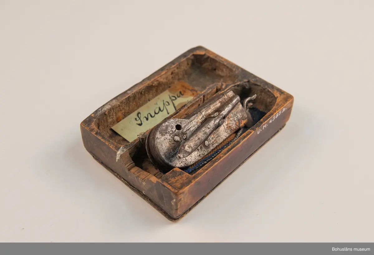 Ur handskrivna katalogen 1957-1958:
Snäppa m. fodral
Snäpparen a) L. 6,5 cm; helt av järn. Hel. Fodralet b) 8,5 x 6,5 x 2 cm; m. fack f. 2 "snäppor"; på undersid. ett lock som skyddar ett spegelglas i 2 delar. Defekt.

Lappkatalog: 94