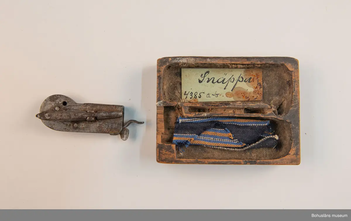 Ur handskrivna katalogen 1957-1958:
Snäppa m. fodral
Snäpparen a) L. 6,5 cm; helt av järn. Hel. Fodralet b) 8,5 x 6,5 x 2 cm; m. fack f. 2 "snäppor"; på undersid. ett lock som skyddar ett spegelglas i 2 delar. Defekt.

Lappkatalog: 94