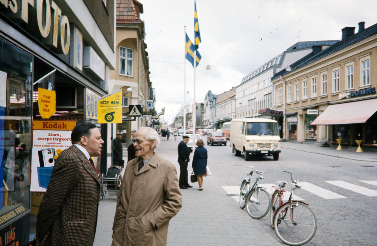 Storgatan, Växjö. Nils Hyltén-Cavallius pratar med en herre på trottoaren. I bakgrunden syns bl a Qvidings bokhandel.
Färgfoto ca 1970.