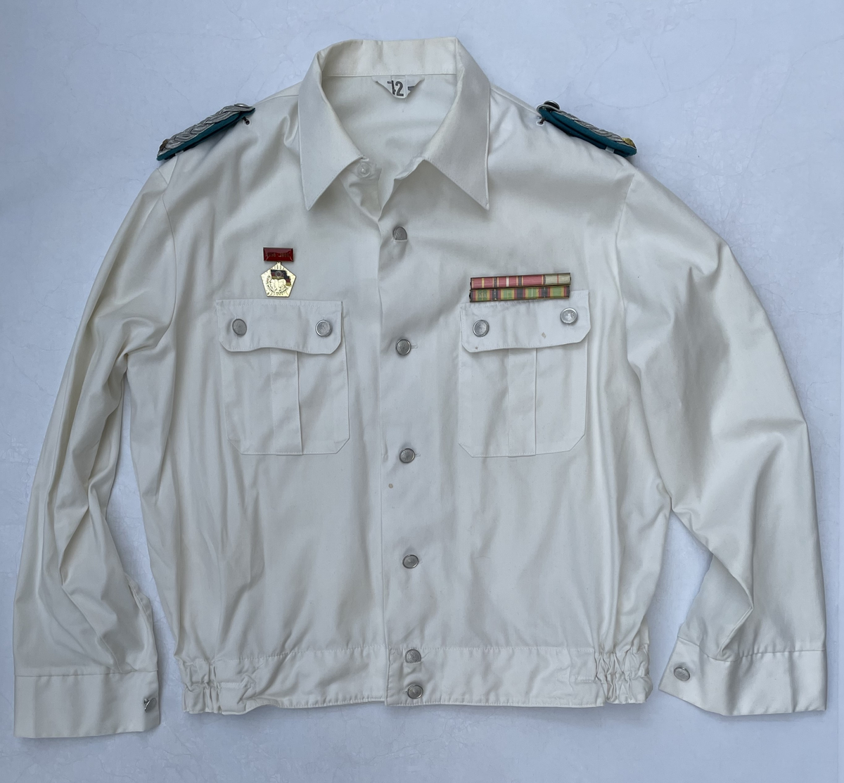 En vit skjortblus (Hemdbluse) för överstelöjtnant vid de östtyska gränstrupperna (Grenztruppen der DDR). Storlek 42N. Skjortblusen ingick i stabsuniform sommar (Stabsdienstuniform Nr. 2 So) och bars utan skjorta under.
Över höger bröstficka sitter en minnesmedalj för östtyska armens (Nationalen Volksarmee – NVA) 30-års jubileum. På nålfästet texten ”1956-1986”, på medaljens framsida ”30 JAHRE NATIONALE VOLKSARMEE” och på frånsidan ”Fest verwurzelt im Volk, gefürt von der Partei, erfüllen wir den Klassenauftrag. Forwärts zom XI. Parteitag der SEDI”.
Över vänster bröstficka sitter släpspännen. 
I övre raden band för (från höger till vänster från betraktarens håll); Vapenbrödramedaljen (Medaille der Waffenbrüderschaft) i brons och östtyska armens förtjänstmedalj (Verdienstmedaille der Nationalen Volksarmee) i brons, silver och guld.
I undre raden band för (från höger till vänster från betraktarens håll); medaljen för trogen tjänst i östtyska gränstrupperna (Medaille für treue Dienste in den Grenztruppen der DDR) 5 års tjänst, 10 års tjänst, 15 års tjänst och 20 års tjänst.