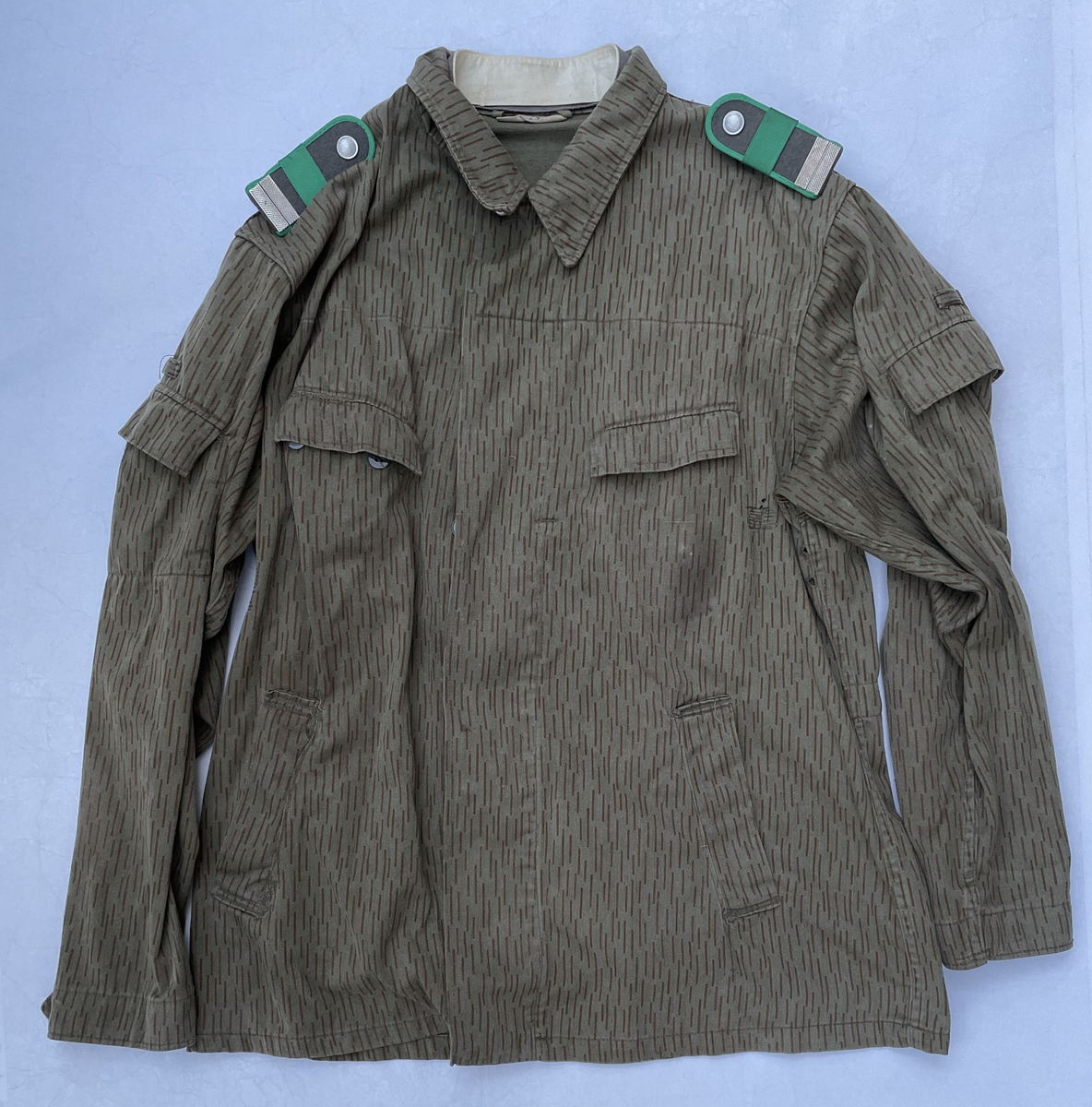 En fältjacka (Feldjacke) för korpral (Stabsgefeiter) och posteringschef (Postenfürer) vid östtyska gränstrupperna (Grenztruppen der DDR). Kamouflage i ”Strichtarn” mönster. Storlek Sg 52-0.
Fältjackan har dolt knäppning. Innerkragen sitter med knappar och går att byta. Jackan har två bröstfickor med dold knäppning under ficklocket, två framfickor och en ficka på vardera överärmen samt en dold innerficka för pistol.
 På insidan, vid kragen, är den märkt ”Hübner” med bläckpenna.